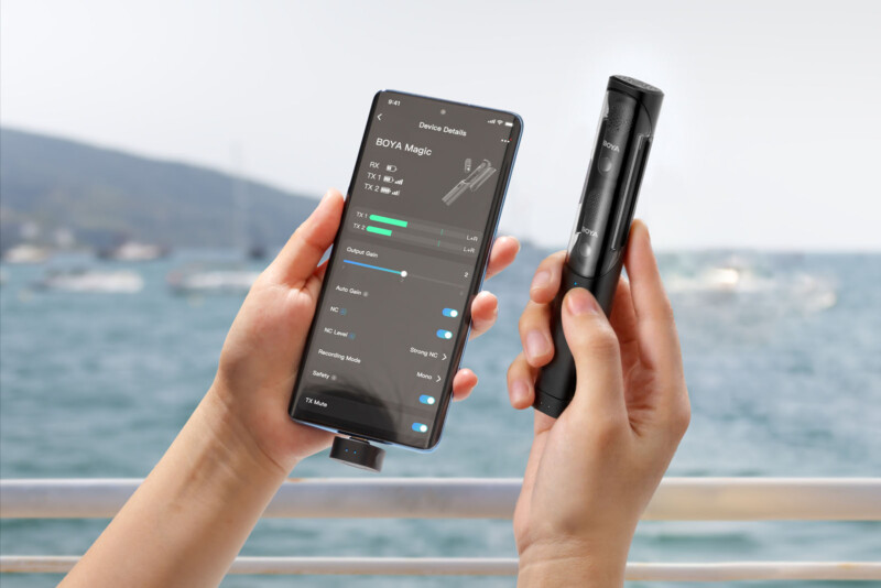 A person holds a smartphone displaying audio device settings in one hand and a black wireless microphone in the other hand, with a blurred seaside background.