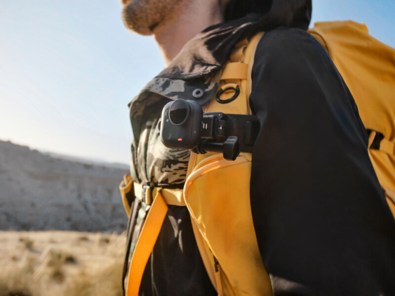 A person wearing a yellow backpack and black jacket has a small action camera attached to the backpack strap, hiking outdoors in a sunny, rocky landscape.