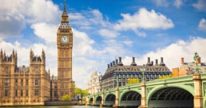 A view of Big Ben and the Houses of Parliament in London, with Westminster Bridge crossing the River Thames under a partly cloudy blue sky.