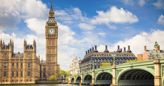 A view of Big Ben and the Houses of Parliament in London, with Westminster Bridge crossing the River Thames under a partly cloudy blue sky.