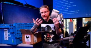 A person with glasses and tattoos holds metal plates while sitting at a desk with a laptop, blue storage bins, and a cardboard box in a brightly lit, tech-filled room. A chat screen is visible in the background.