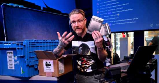 A person with glasses and tattoos holds metal plates while sitting at a desk with a laptop, blue storage bins, and a cardboard box in a brightly lit, tech-filled room. A chat screen is visible in the background.
