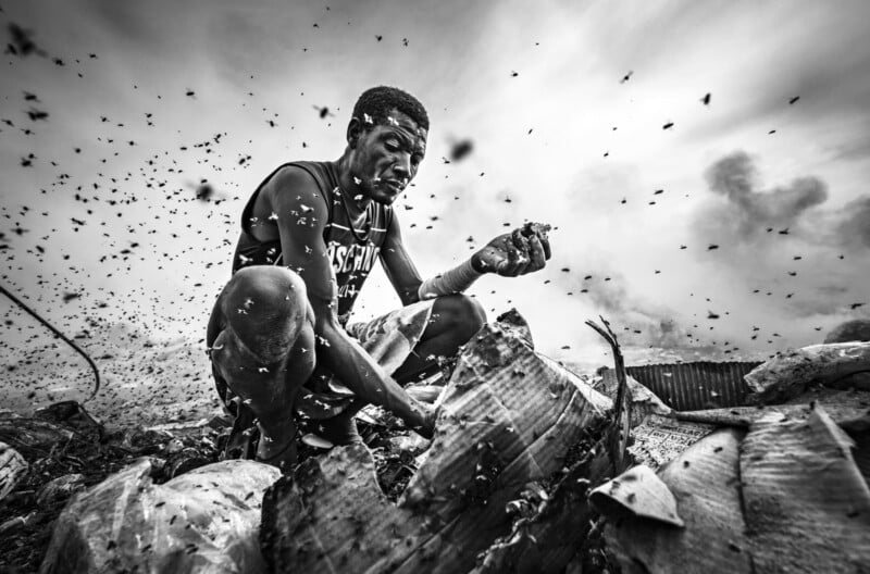 A man crouches amidst piles of debris and trash, surrounded by swarming flies. The scene is chaotic, with smoke in the background and a cloudy sky above, highlighting harsh living conditions. The image is in black and white.