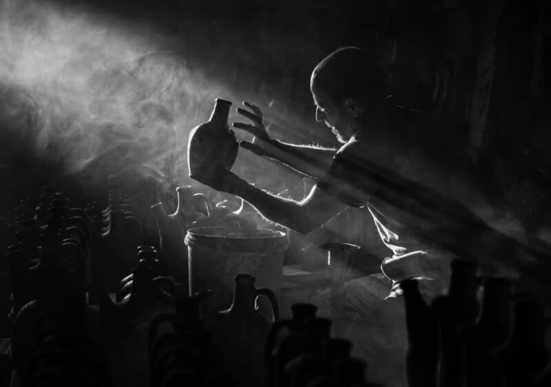 A person in a dark workshop holds a clay jug up to the light, surrounded by pottery and illuminated by rays of sunlight filtering through swirling dust and smoke.