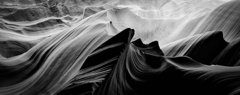 Black and white photo of smooth, curving rock formations with dramatic shadows and textures, resembling waves or flowing fabric in a canyon. Light highlights the ridges and depth of the landscape.