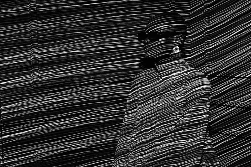 A person wearing sunglasses stands against a wall covered in horizontal, wavy, black-and-white lines that also cast dramatic, striped shadows across their face and clothes. The overall effect is abstract and visually striking.