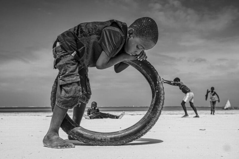 A boy leans his body over a large tire on a sandy beach while other children play in the background under a partly cloudy sky. The scene is captured in black and white.
