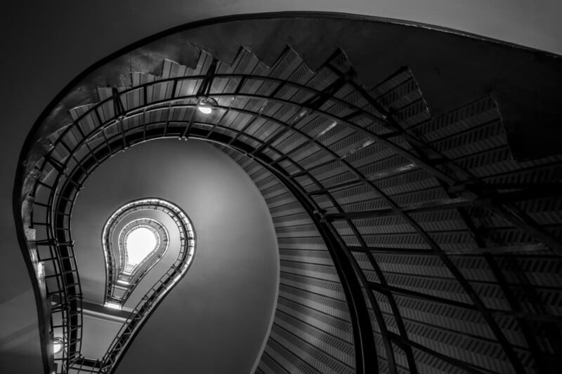 Black and white photo of a spiral staircase taken from below, forming the shape of a light bulb with a glowing light at the top center, and detailed railings and steps curving upward.