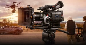 A professional film camera is set up outdoors near a road, with a convertible car, a drone flying overhead, and two crew members standing nearby. The scene appears to be part of a film production in a desert area.