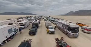 Rows of RVs, campers, and cars line up on a wide, flat desert under a cloudy sky, with mountains in the distance and people moving around the vehicles.