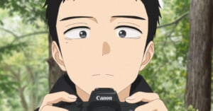 An anime-style character with short black hair holds a Canon camera, looking slightly upward with a neutral expression. The background shows green trees and a forest setting.