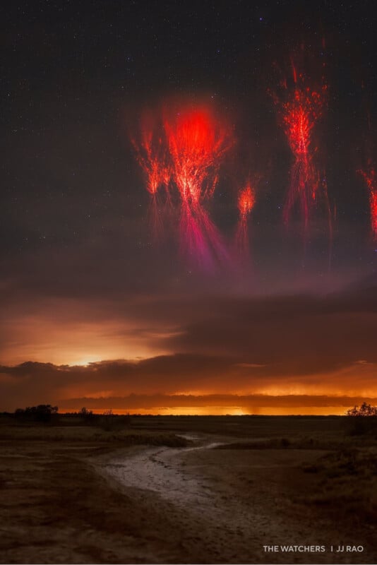 A night sky glows with red sprite lightning above distant clouds, illuminating the landscape below with an orange hue. A winding path leads through the dark and barren foreground.