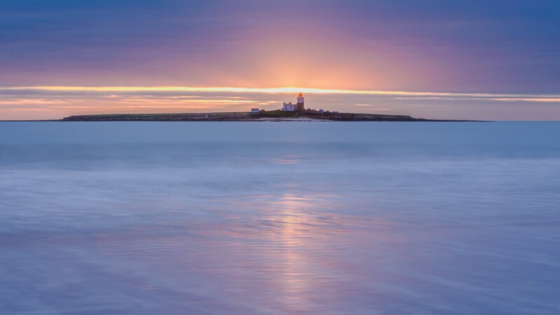 A lighthouse on a small island is illuminated by the soft glow of a sunset, with calm sea waves in the foreground and a colorful sky with purple and orange hues in the background.