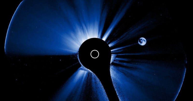A blue-tinted image of the Sun is partially obscured by a circular black disk in the center, while a small, distant Earth appears on the right side of the image against the blackness of space.