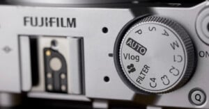Close-up of a Fujifilm camera, showing the mode dial with options like AUTO, Vlog, FILTER, and custom settings. The Fujifilm logo and a hot shoe mount are also visible.