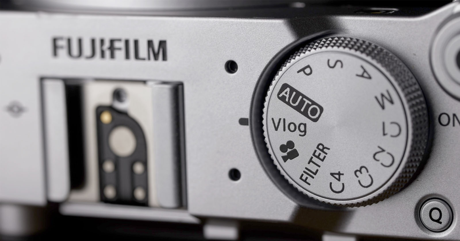 Close-up of a Fujifilm camera, showing the mode dial with options like AUTO, Vlog, FILTER, and custom settings. The Fujifilm logo and a hot shoe mount are also visible.