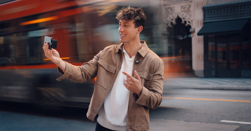 Un joven con cabello rizado sonríe y señala la cámara o el teléfono mientras toma una foto en una calle de la ciudad, pasando por un autobús borrado en el fondo.