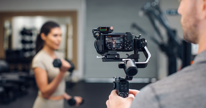 Una persona está disparando a una mujer que levanta las pesas en el gimnasio con una cámara profesional montada en un estabilizador. La mujer está un poco borrosa en el fondo, mientras que la cámara y su monitor están enfocados.