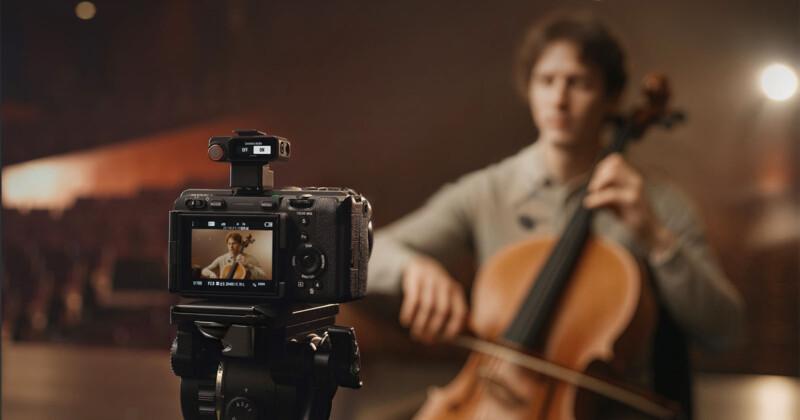 La cámara digital en el trípode graba el músico tocando violonchelo en el escenario. El monitor de la cámara muestra el enfoque del músico mientras el fondo y el músico parecen borrosos. La atención brilla en el Auditorio Dim.