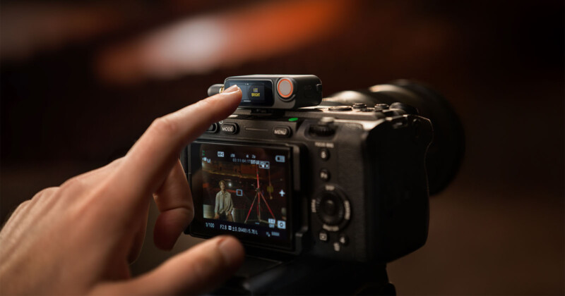 Una persona ajusta un pequeño dispositivo que está montado en la parte superior de una cámara digital, mientras que la pantalla de la cámara muestra que la escena se está filmando, incluida la persona que se encuentra frente a una habitación cálida frente a un trípode.