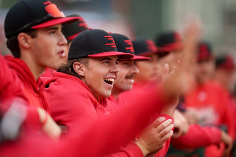 A group of baseball players in red uniforms and black caps sit together, smiling and cheering enthusiastically at the game. The focus is on one player in the center, who is laughing and clapping.