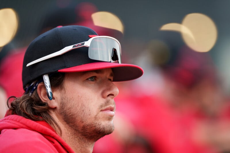 A baseball player with long hair wearing a red hoodie, black and red cap, and sunglasses perched on the cap, looks focused. The background is blurred with other people in red and circular lights.