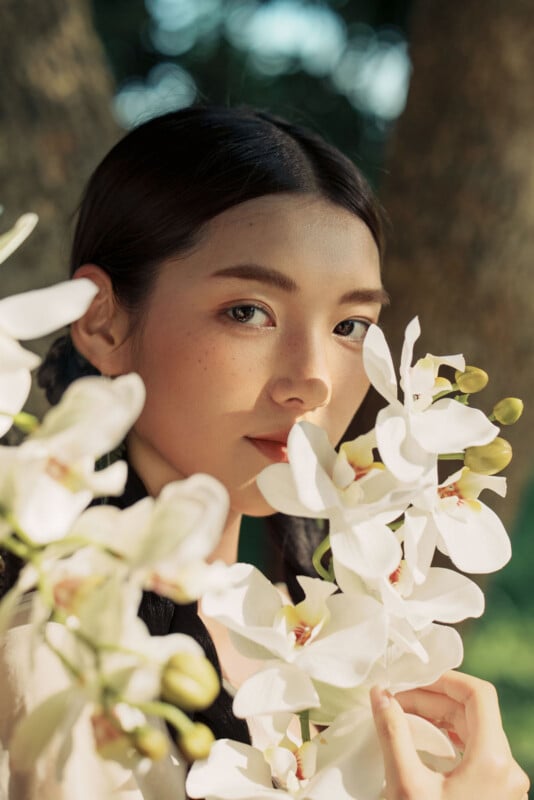 A young woman with long dark hair looks at the camera while holding a cluster of white orchids near her face, with soft sunlight and trees in the background.