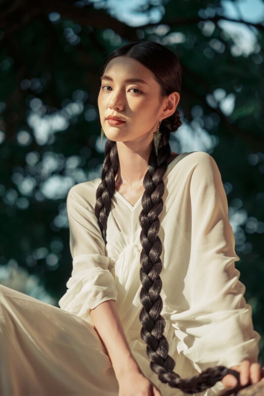 A woman with very long, dark braided hair wears a flowing white outfit. She sits outdoors with sunlight casting shadows on her face and trees blurred in the background.