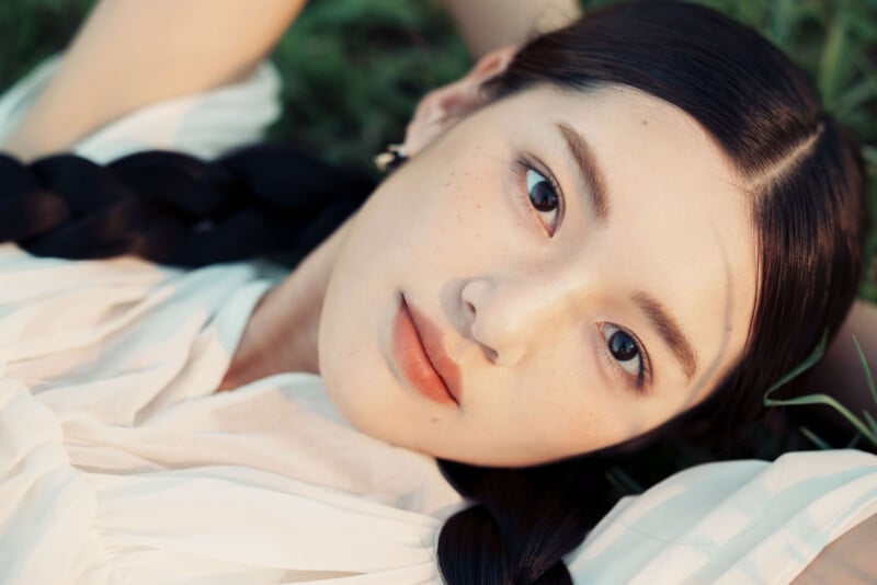 A young woman with long, dark braided hair lies on the grass, gazing calmly at the camera. She wears a light, flowing top and natural makeup, with sunlight softly illuminating her face.