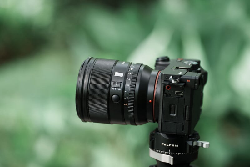 A black digital camera with a large lens is mounted on a tripod, set against a blurred green background of foliage.