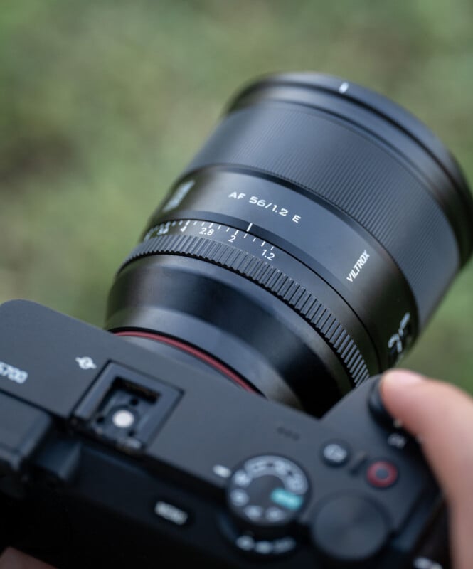 A close-up of a person holding a digital camera with a Viltrox AF 56mm f/1.2 E lens, showing the camera’s controls and lens markings, with a blurred green background.