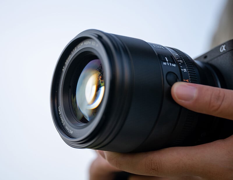 Close-up of a person’s hands holding a black digital camera with a large lens, pointed slightly upward, against a blurred, light background.