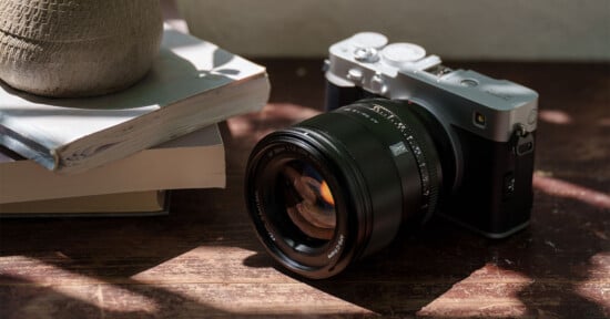 A digital camera with a large lens rests on a wooden surface next to a stack of books and a textured pot, with dappled sunlight casting shadows across the scene.