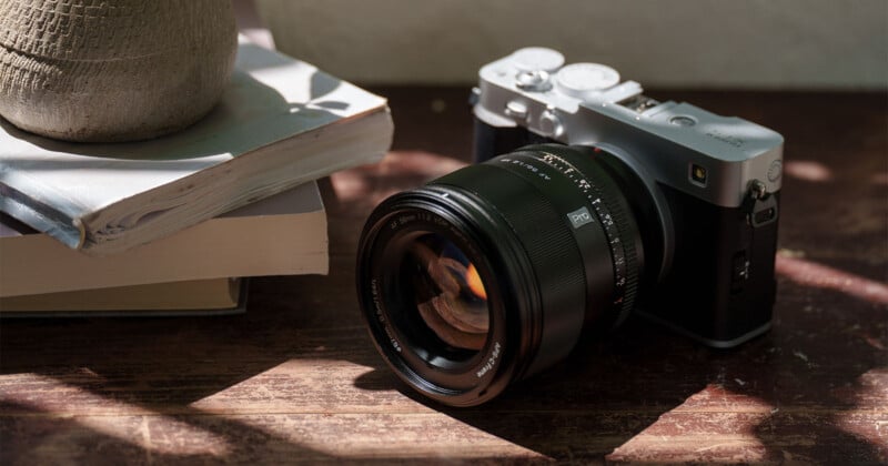 A digital camera with a large lens rests on a wooden surface next to a stack of books and a textured pot, with dappled sunlight casting shadows across the scene.