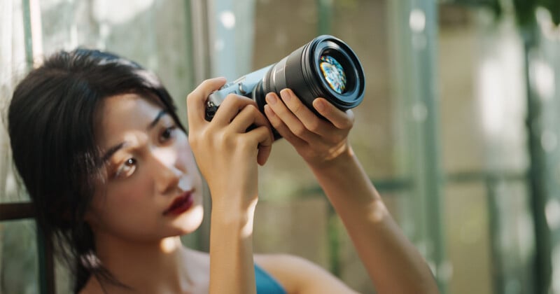 A woman with long dark hair holds a camera up, adjusting the lens and looking intently at it. Soft sunlight filters through a window, illuminating her face and the background.