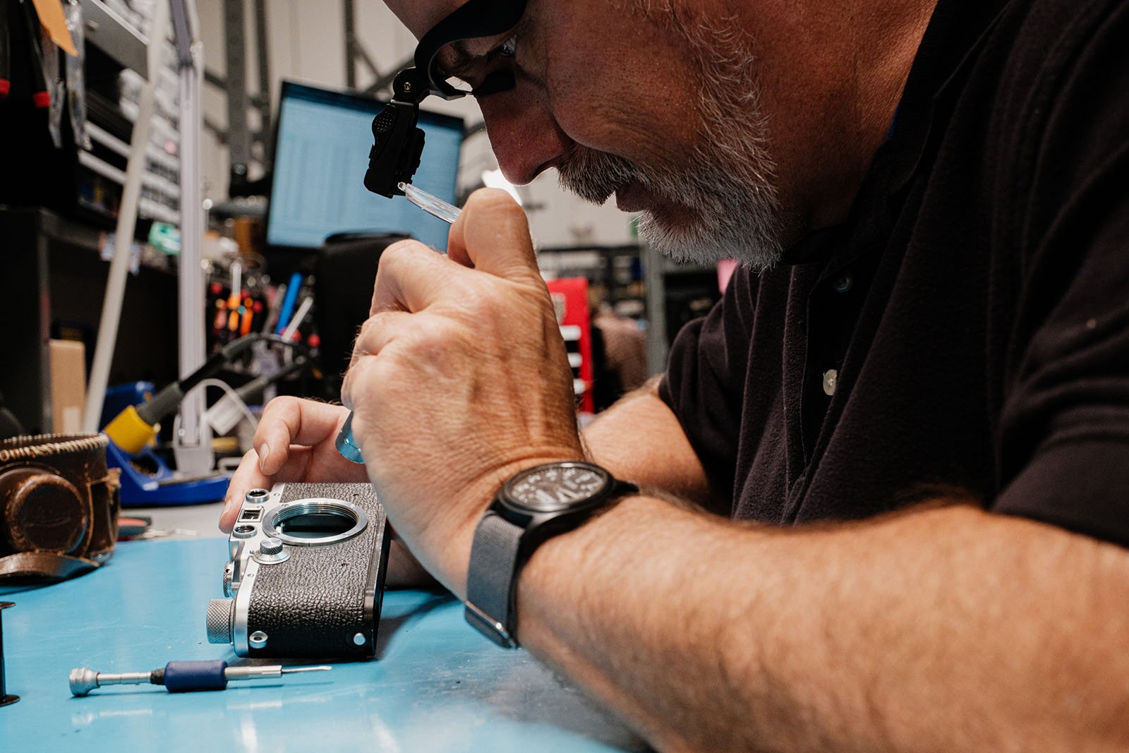 A person wearing glasses and a watch carefully repairs a vintage camera on a blue workbench, surrounded by small tools and electronic equipment.
