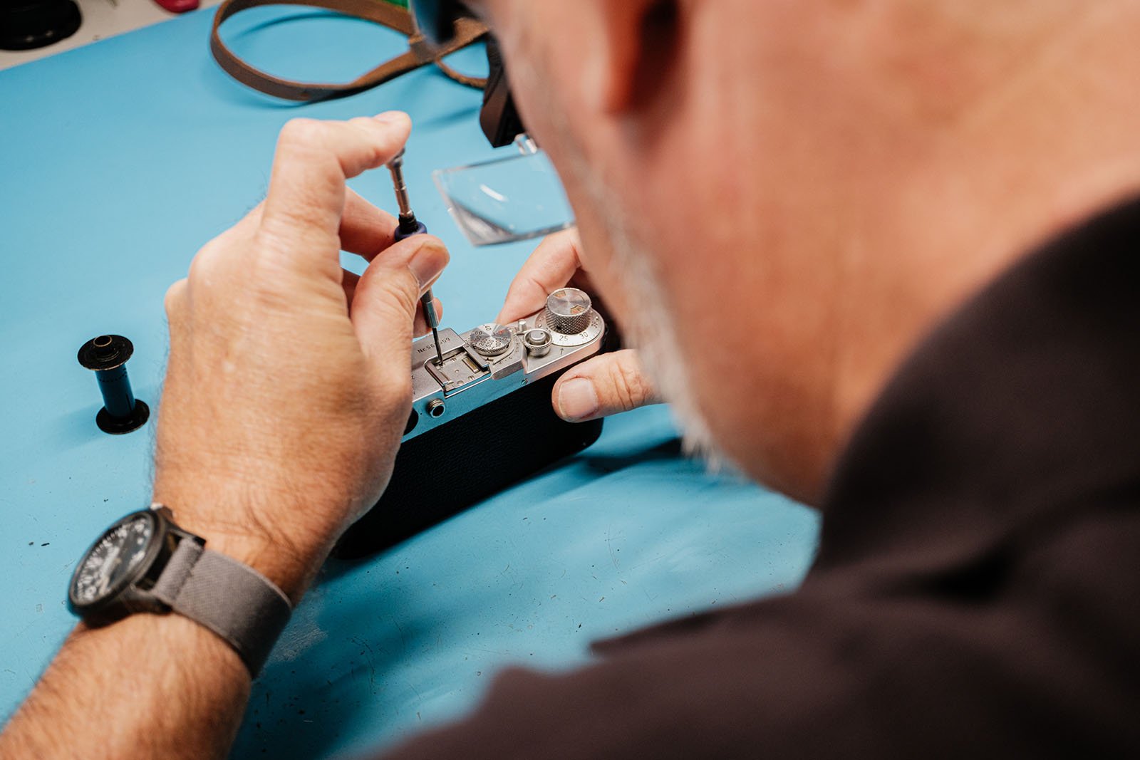 A person wearing a watch uses a small screwdriver to repair or adjust a vintage film camera on a blue surface.