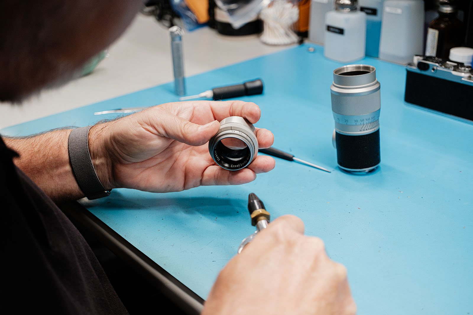 A person holds a camera lens above a blue work surface, with tools and another lens nearby, preparing to work on or repair the lens.