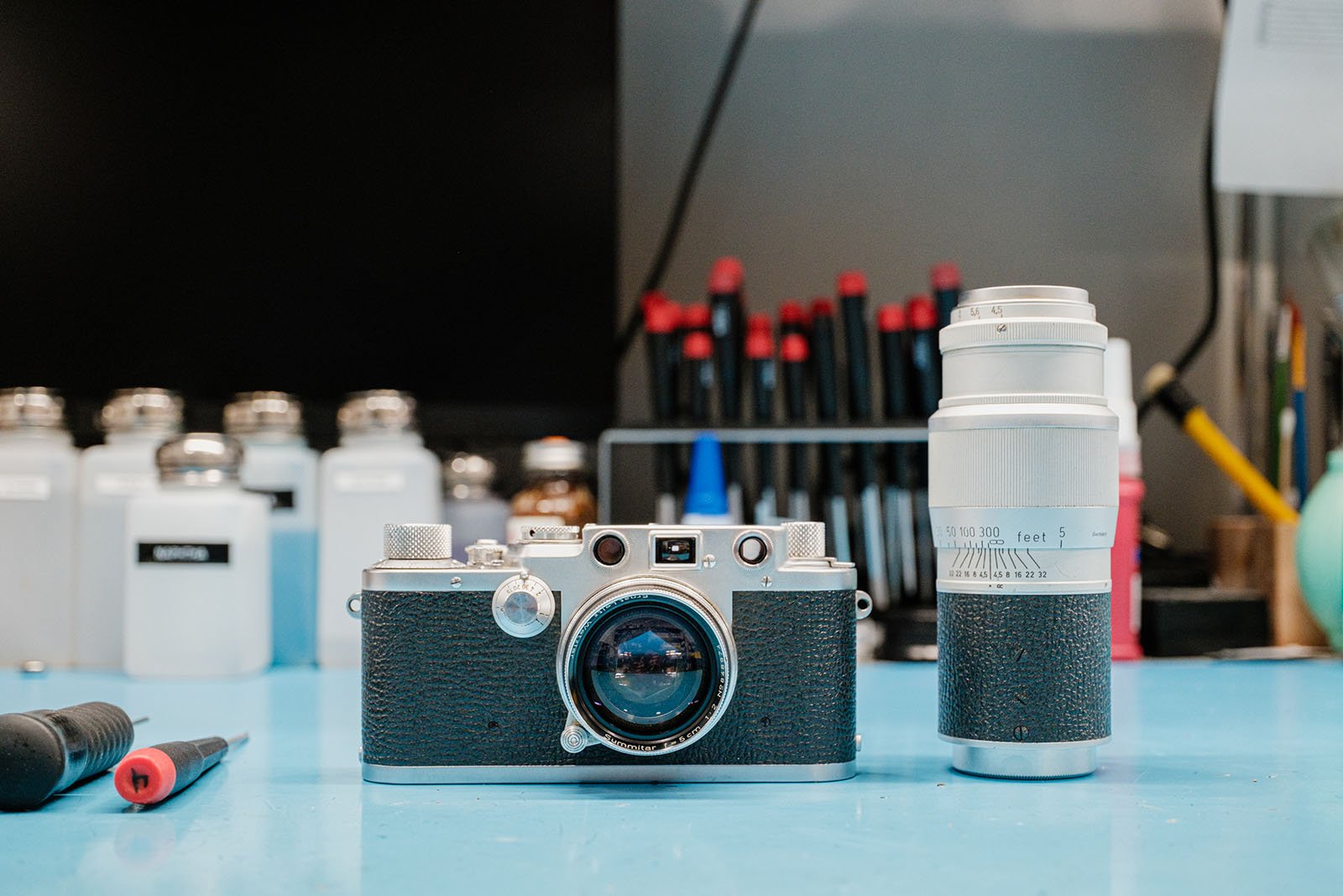A vintage camera and a silver telephoto lens are placed on a blue workbench, with tools, jars, and various equipment in the background.