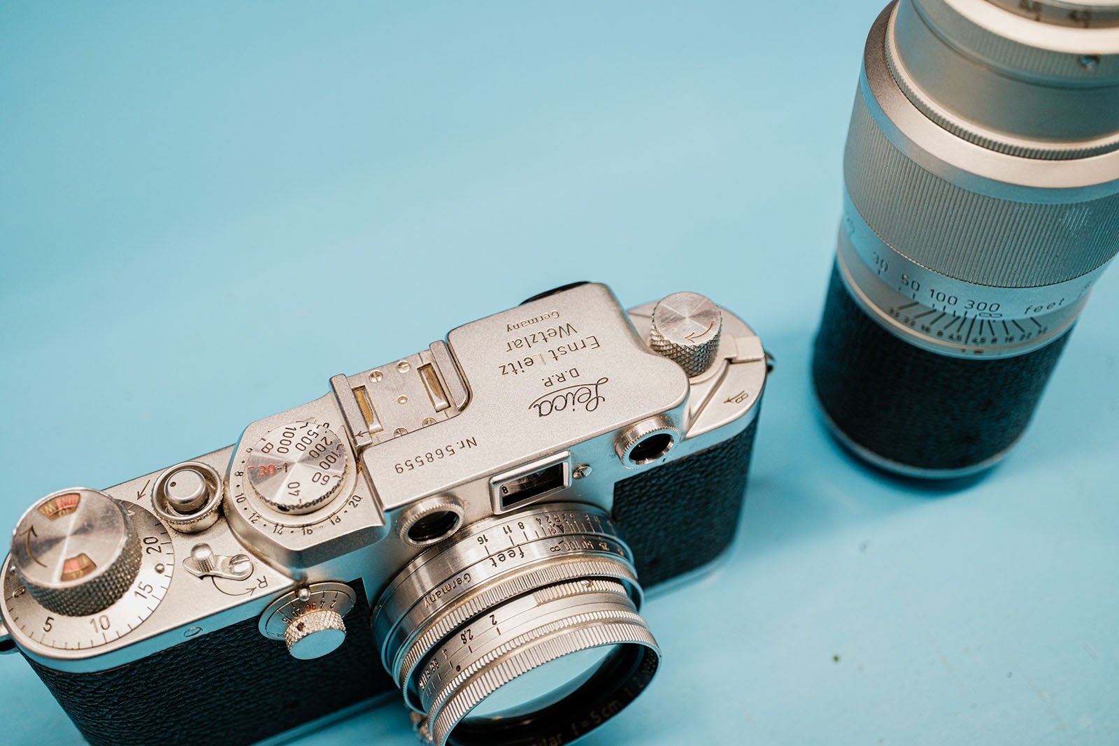 A vintage Leica camera and a separate telephoto lens are placed on a light blue surface. The camera has a metal body with engraved text and a black textured grip.