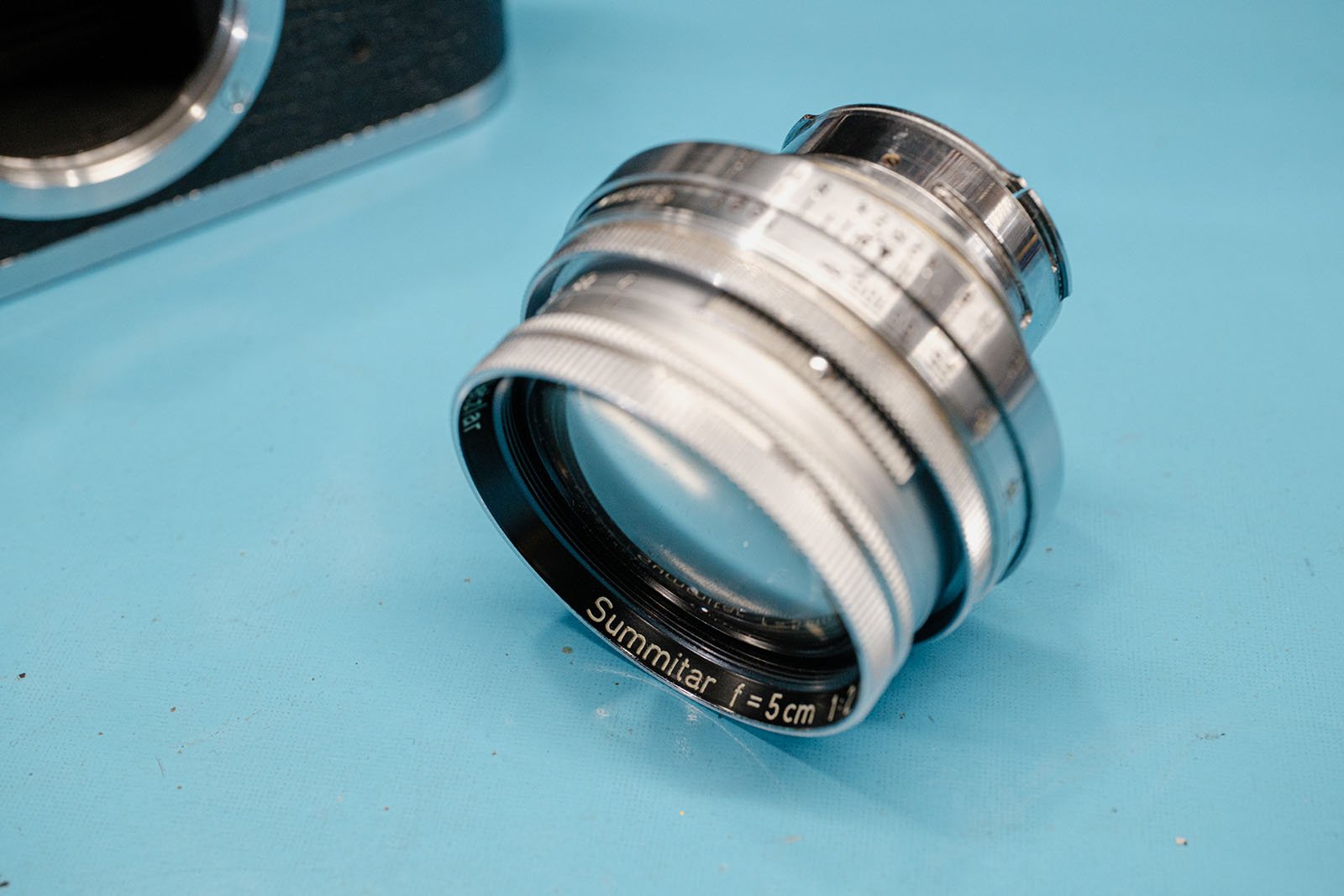 A vintage Summitar 5cm camera lens with silver metal body is lying on a blue surface beside part of a camera body. The lens markings and glass elements are visible.
