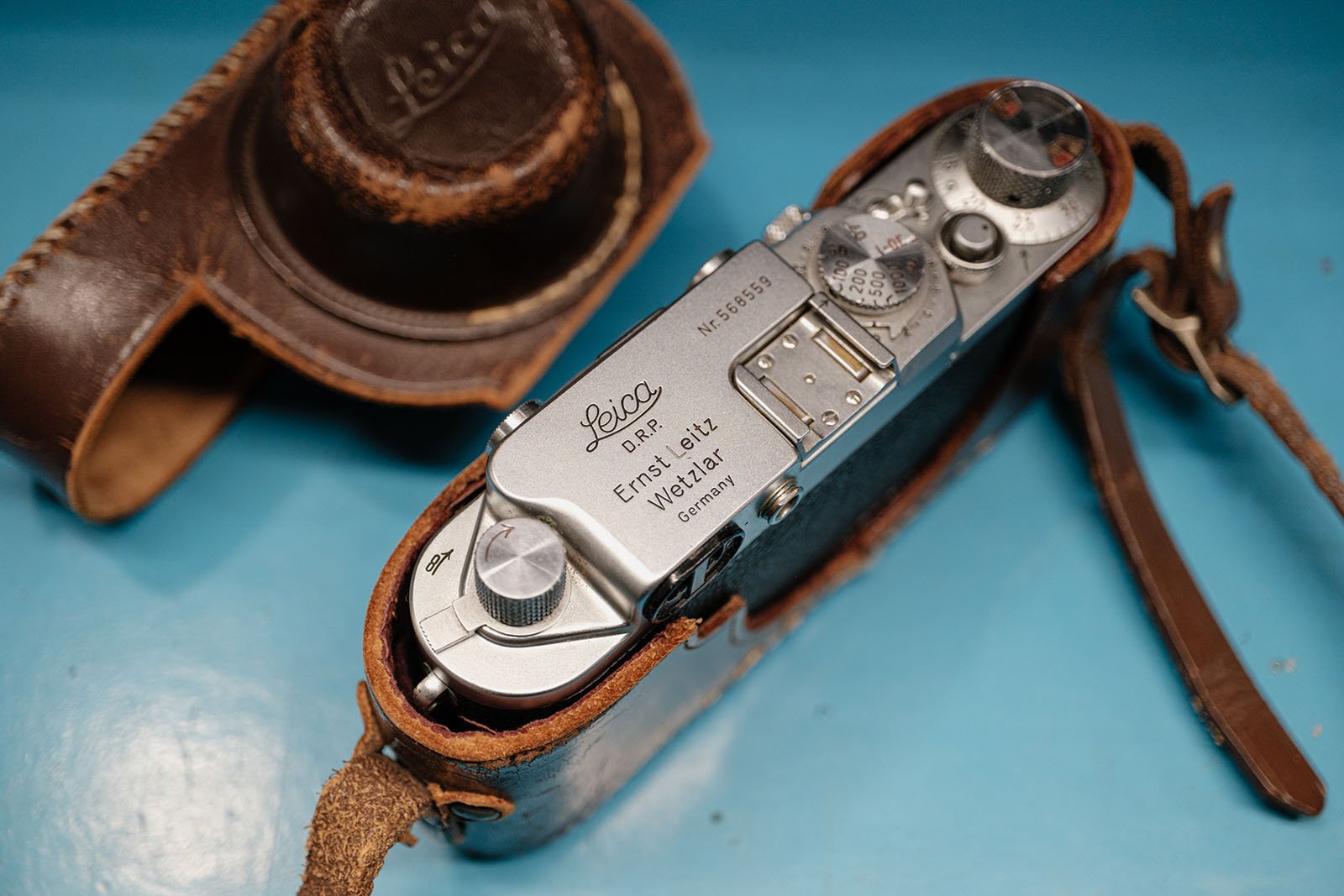 A vintage silver Leica camera in a brown leather case, with engraved text and dials visible on top, rests on a blue surface. The open case lid is positioned nearby.
