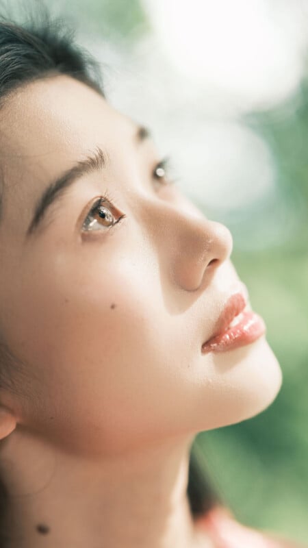 A close-up of a young woman’s face looking upwards, with soft natural light highlighting her smooth skin and reflective eyes. The background is blurred with shades of green and white.