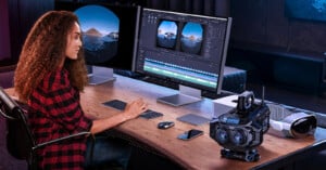 A woman sits at a desk editing 360-degree video footage on dual monitors. The desk has a VR headset, a futuristic camera, a smartphone, a keyboard, and a mouse. The workspace is modern and well-lit.
