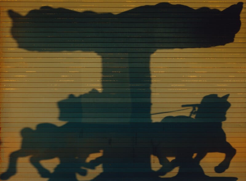 Shadow of a carousel with two horses is cast on a yellow metal wall, creating a silhouette effect with strong outlines of the horses and carousel roof.