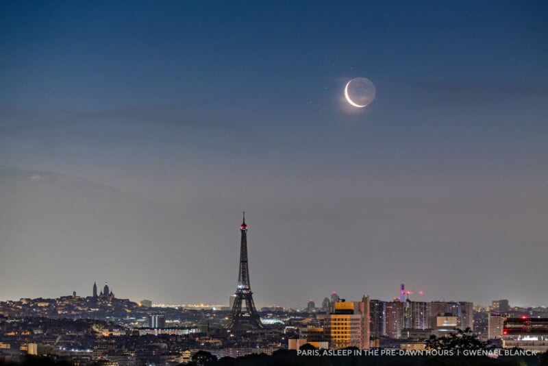Pre-dawn view of Paris with city lights glowing, the Eiffel Tower at center, and a crescent moon with earthshine visible in the clear sky above.