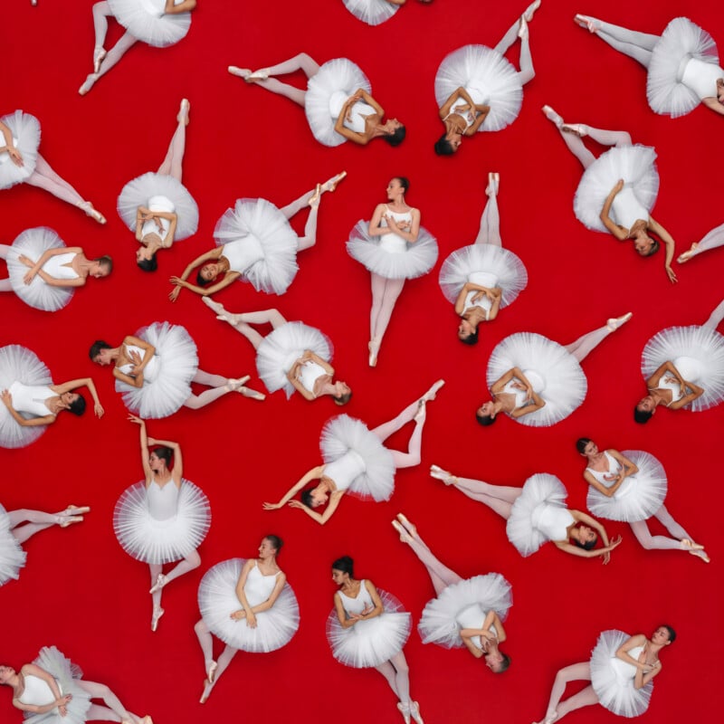 Ballet dancers in white tutus pose gracefully on a bright red background, viewed from above. The dancers are arranged in various positions, creating a striking and elegant pattern.
