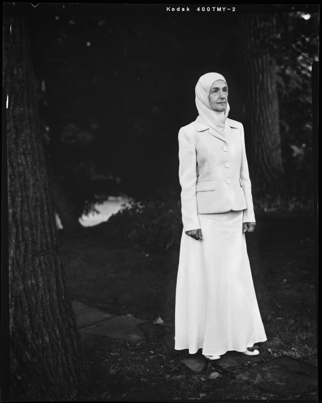 A woman wearing a light-colored hijab, blazer, and long skirt stands outdoors on a stone path, surrounded by trees and foliage. The black-and-white photo has a tranquil and contemplative atmosphere.