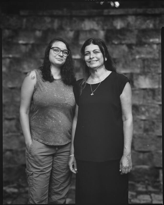Two women stand closely together in front of a stone wall. One wears glasses, a sleeveless shirt, and cargo pants; the other wears a short-sleeve dress. Both are looking at the camera and smiling slightly. The photo is black and white.