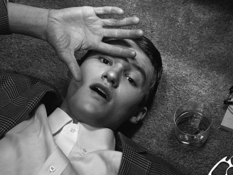 A young man in a suit lies on a carpeted floor, looking up with a surprised expression, one hand raised to his forehead. A tumbler glass with liquid sits nearby. The image is in black and white.
